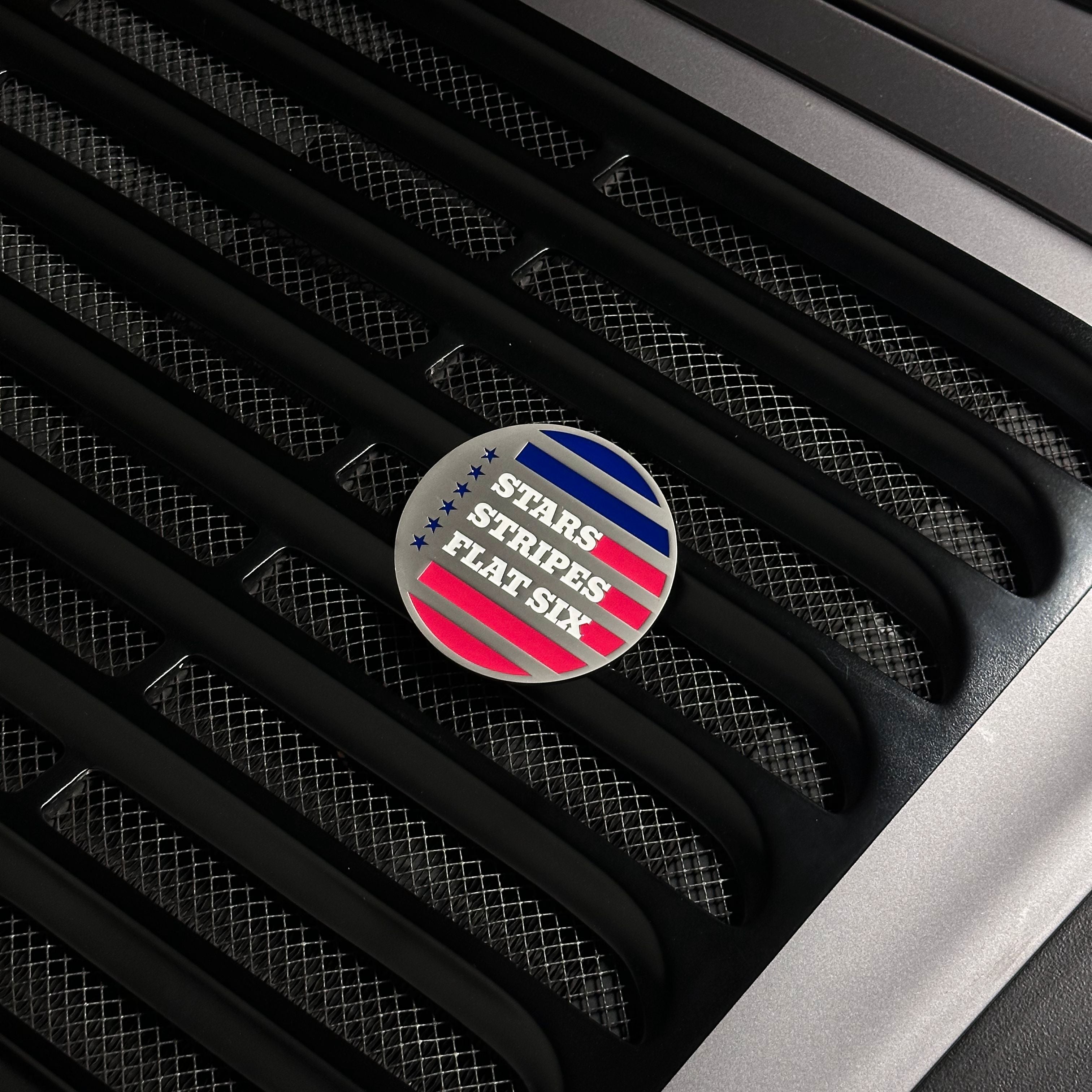Round badge with 'Stars Stripes Flat Six' design on a car grille.